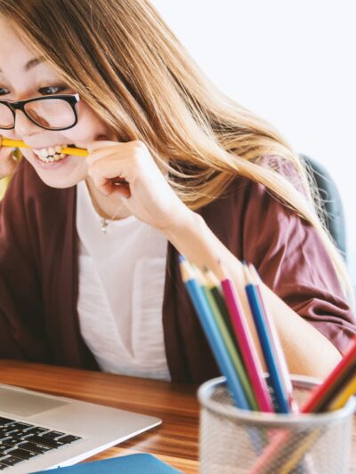 woman biting pencil while sitting on chair in front of computer during daytime